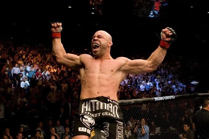 Bald male fighter celebrating in octagon with arms raised, surrounded by crowd in arena during MMA event