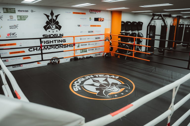 Professional boxing ring with black mat and logo at Shogun Fighting Championship gym, featuring orange and white railings and training equipment